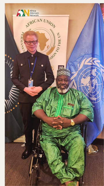Two men posing together in an office setting. On the left, a standing man with albinism wears glasses, a black suit jacket, black trousers, and a UN lanyard badge. On the right, a man with a grey beard sits in a wheelchair wearing a green patterned traditional outfit and matching cap. Behind them are banners for the African Union Permanent Observer Mission to the UN and a large blue United Nations flag.