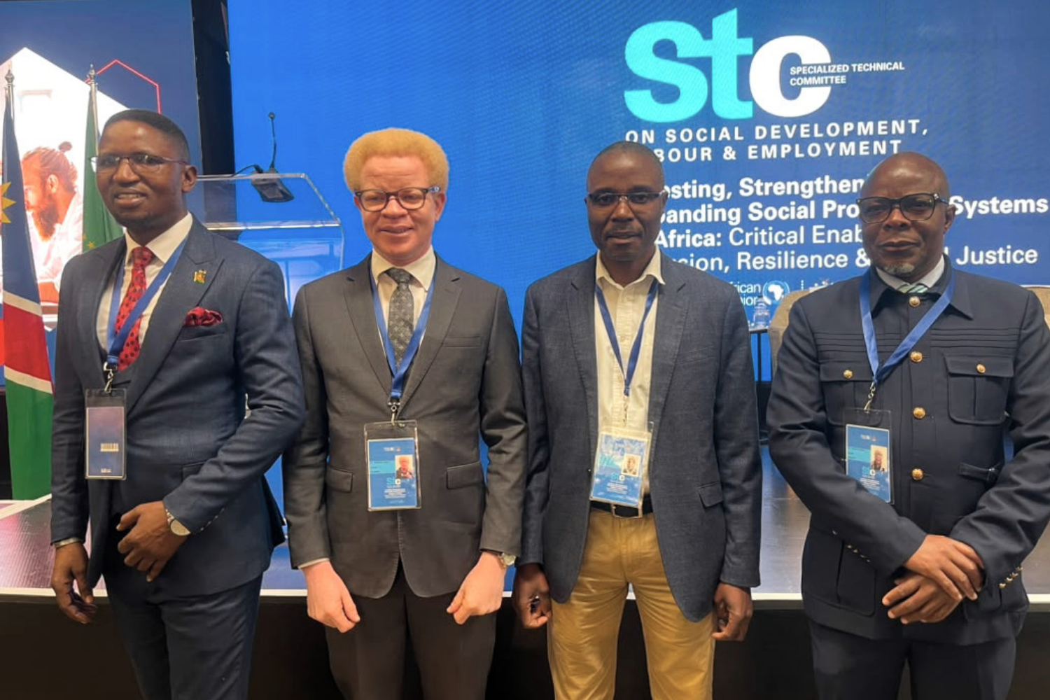 Four male delegates wearing official conference credentials stand side-by-side in a brightly lit indoor venue. In the background, a large screen displays the logo for the Specialized Technical Committee (STC) on Social Development, Labour & Employment along with theme text regarding social protection systems.